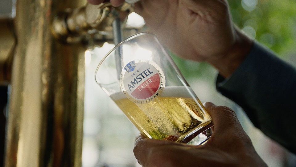 Fotografia de uma mão servindo um copo de chopp Amstel.